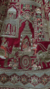 Close-up of royal procession embroidery with camel riders and architectural details on a red silk bridal lehenga.
